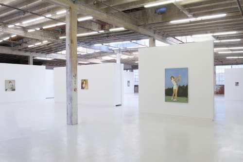 Hosfelt Gallery, looking at the west gallery. All of the floors and walls are painted white. a square shaped wall has a large rectangular painting, depicting a woman having just swung a golf club. Another floating wall to the left has a small square paint