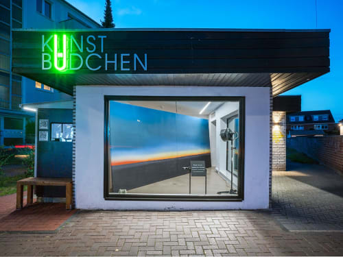 glass-front booth with neon letters spelling 'KUNSTBÜDCHEN' holds a large landscape canvas within, wedged sideways
