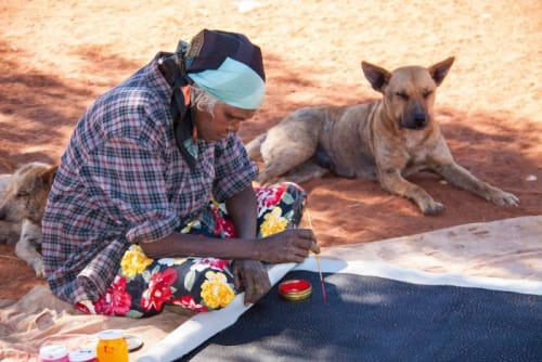 a woman sitting in dirt, with a canvas rolled out in front of her. she is using red paint to dot her canvas. She is wearing a blue plaid shirt and floral pants, with a bandana on her head. there is a wile dog laying next to her.
