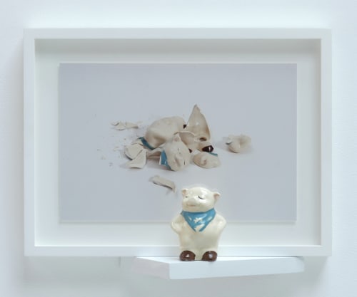 A wall installation of a ceramic pig mounted in front of a framed photograph of the same ceramic pig shattered on the ground