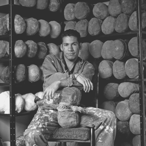 Rafael Gomez Barros artist photo. He sits in his studio, wearing overalls covered in paint and plaster. He is backgrounded by works from his "Sonjaeros" (rattles) series. The sit on shelves behind his except for one which sits between his legs, he is rest