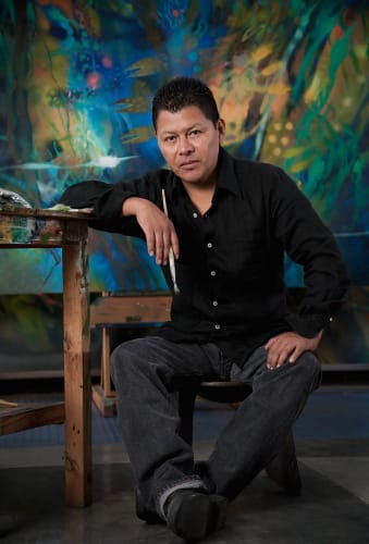 Carlos Jacanamijoy artist photo. He sits in his studio, left arm resting on a table, holding a brush. He is backgrounded by one of his works.