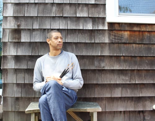 Artist Mario Andres Robinson sitting on a bench holding paintbrushes against a sidewall of a wooden house