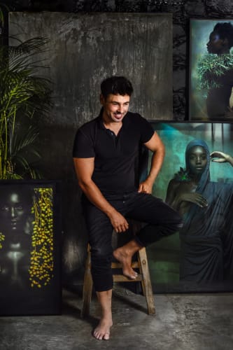 Artist Krisjan Rossouw in black and barefooted, sitting on a high stool looking down away from the camera, having his photographs in the background