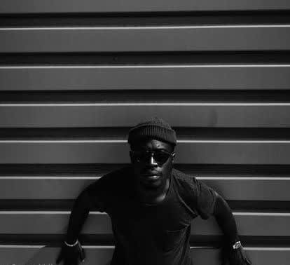 Artist Malick Kebe in a monochromatic photography against a wall wearing sunglasses and a hat