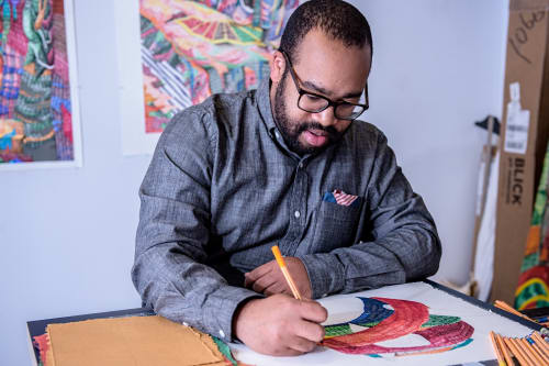 Artist Michael Andrew Booker at the desk of his studio working on his painting, having another two in the background.