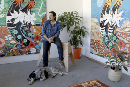 Daniel Gibson seated in his studio with his dog resting on the floor in front of him and two of his paintings hanging on the walls behind him