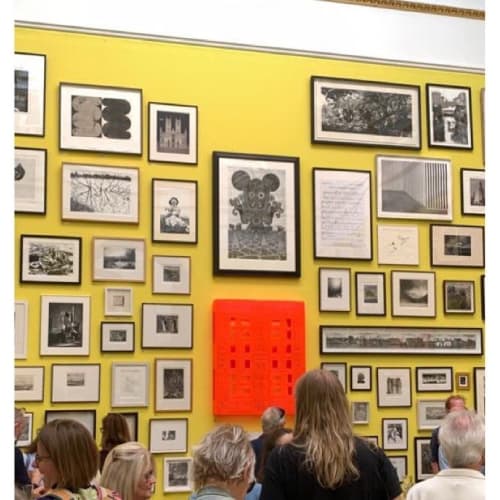 An image of a gallery at the Royal Academy Summer exhibition. In the centre is Ade Adesina's print 'Parallell' surrounded by numerous other monochrome works. They are installed on lemon yellow wall. A crowd observes the works from below