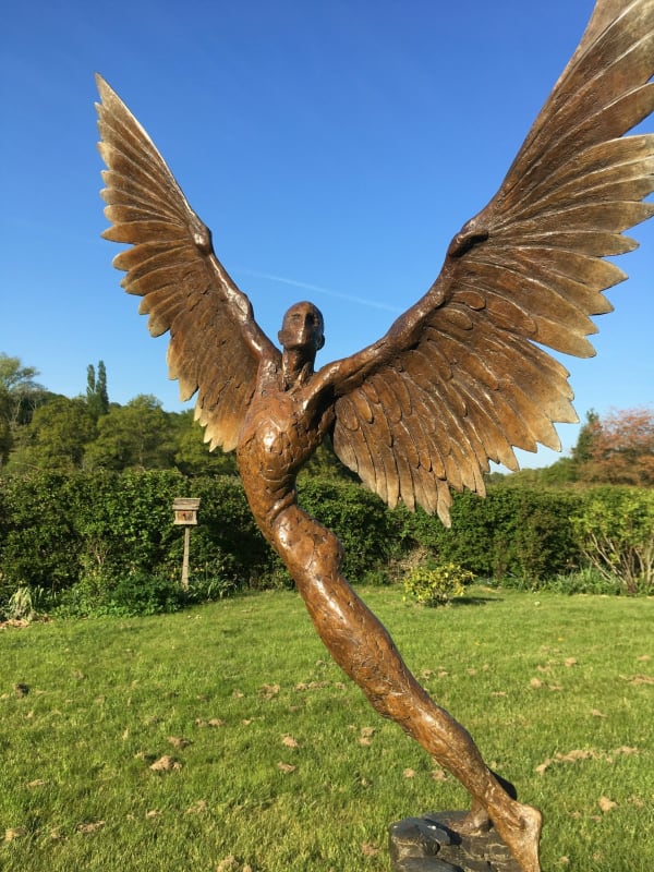 a bronze mythological winged figure, on grass in front of a hedge