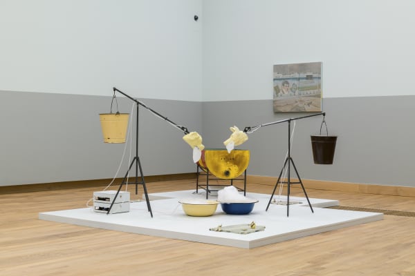 exhibition view of an installation consisting of two ceramic dog heads at the end of tripod stands. these figures are both balanced by buckets. the ceramic heads foam at the mouth, the foam then falls into large dishes which are fed back into the heads through pumps
