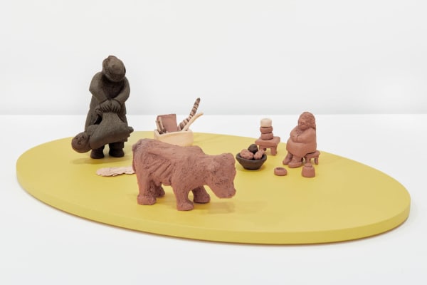 very small ceramics pieces atop a yellow platform. the pieces depict a person holding a bag containing a torso, a basket of objects, a cow, a bowl of what appear to be potatoes, and a very small sitting figure