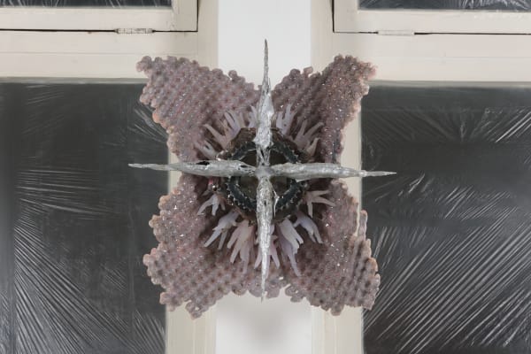 An image of a sculpture consisting of a star shaped metal structure framed by pink pigmented silicone, hanging on a white wall in between two windows blocked off by wrinkled black plastic.