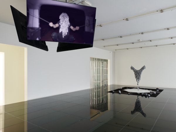 An image of a room with white walls and black marble floor; in the foreground are television screens displaying an image of a masked figure pulling ropes with both arms and in the background is a silver v-shaped structure standing in the center of a black and white square mat.