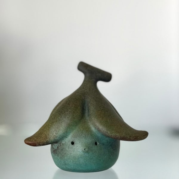Bronze sculpture of a small round figure with a peeled banana on its head in a gradient brown to turquoise green