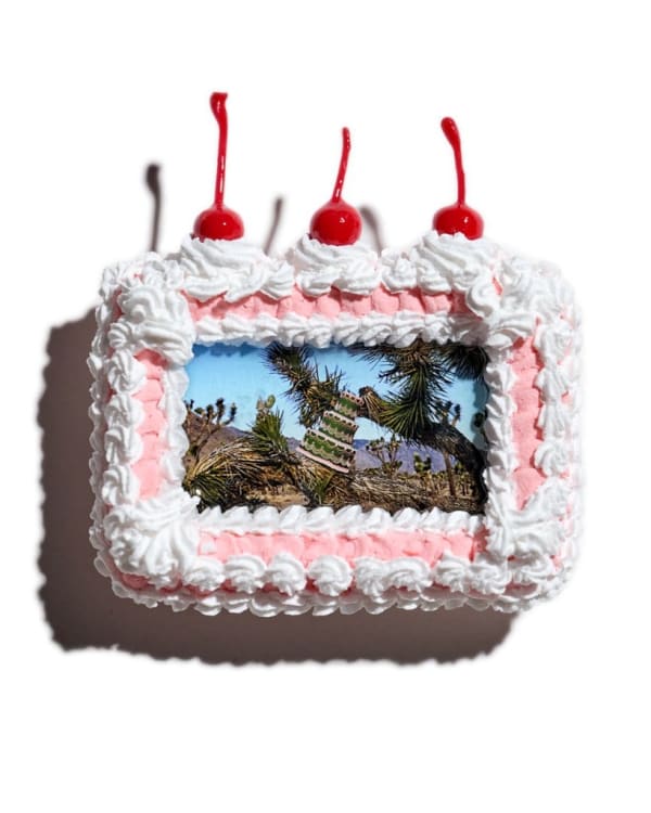 A picture frame that looks like a pink and white cake with three cherries on top. Inside the frame is a 3 layered cake surrounded by joshua trees.