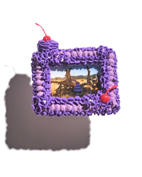 A picture frame that looks like a purple cake with two cherries. Inside the picture frame is a photo of a three layer purple cake in the middle of the desert.