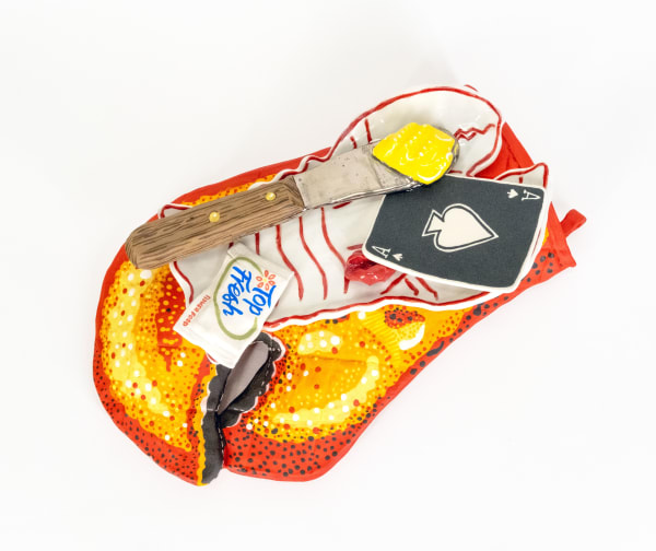 Ceramic sculpture of a lobster claw shaped oven mitten with a lobster shaped plate on top of it. On top of the plate is a knife with butter, an ace of spades playing card and a wet wipe.