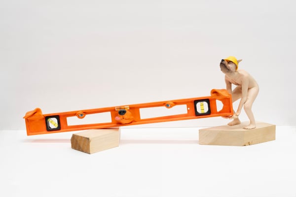 sculpture of nude miniature anthropomorphic dog wear a construction hat and struggling to pick up a level tool