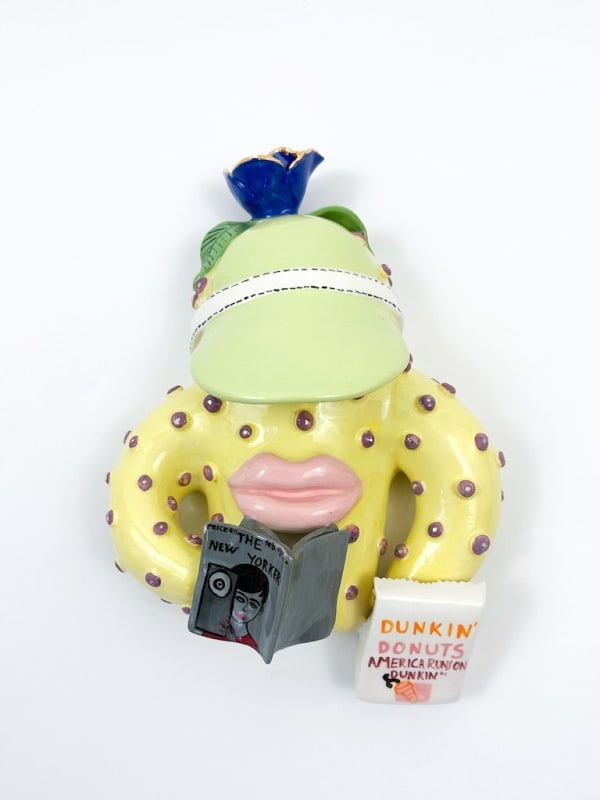 Ceramic creature with large lips holding a book and a Dunkin Donuts bag and wearing a green visor