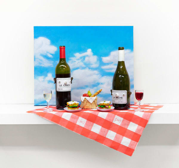 Installation featuring two bottles of wine with dog faces, two sandwiches with dog faces, a picnic basket with a baguette, an apple and a pear all with dog faces on top of a red and white checkered table clothe and a painted sky with clouds in the shape of dogs.