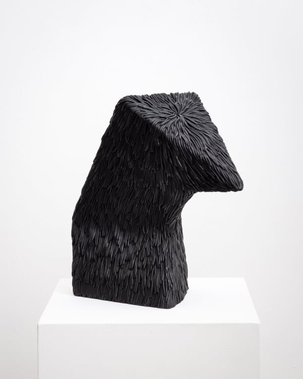 Free-standing bronze sculpture of an abstracted form with a flat triangular head that is bowing down. The sculpture is cast with a surface texture resembling shaggy hair and treated with a black patina.