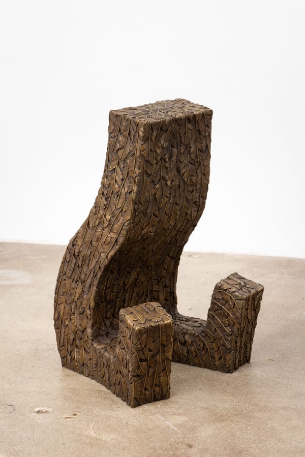 Free-standing bronze sculpture of a rectangular form sitting with outstretched legs. The sculpture is cast in surface texture resembling feathers and treated with a golden brown patina.