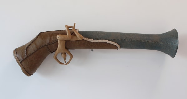 Cardboard and paper mache sculpture takes the appearance of stitched leather Blunderbuss gun At the end of the hun a humanoid figure takes the form of the the trigger and on the inside of the gun’s bell another humanoid lies cradled in a fetus position