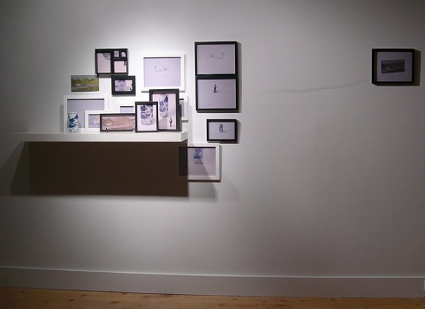 Digital photograph of an installation in which a white wooden shelf is surrounded by multiple variously sized picture frames. The picture frames contain photograms of figurines, drawings, and paintings and some are connected to each other by a string. On the white shelf a tiny human figurine and a tall white porcelain vase are placed. These two figurines are the same ones displayed in the framed photos