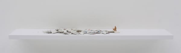 Digital photograph of an installation piece in which multiple pieces of broken ceramic shards are arranged in a horizontal pile. The ceramic pieces are placed on top of a white wooden shelf which is mounted onto a white wall. Towards the right of the piece a small metallic golden figurine with a watering can stands above some of the ceramic shards