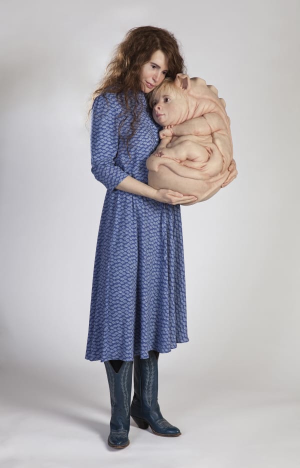 Sculpture of woman in blue dress and boots holding hybrid creature. Creature has human skin, pig ears, and a geometric scaled back. It has many human hands and feet and is curled up in the woman’s arms.