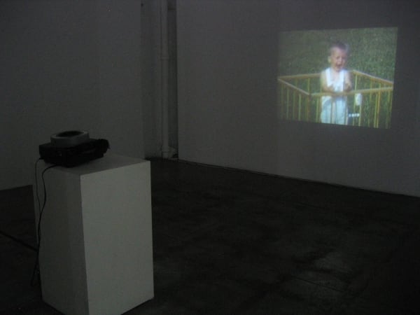 Still image of a projection of a collection of old family home movies onto a wall with slide projector on a pedestal in the foreground to the left