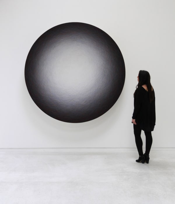 Layers of multi-colored threads strung across a round wooden and plaster panel create the effect of a light gray glowing center that is encircled by a dark gray and black border To the right of the piece there is a woman present to demonstrate scale