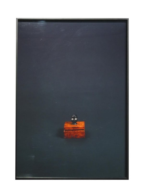 Photographic print displaying a small brown wooden box with a tiny round black robotic head on top. The two figurines are photographed against a dark background