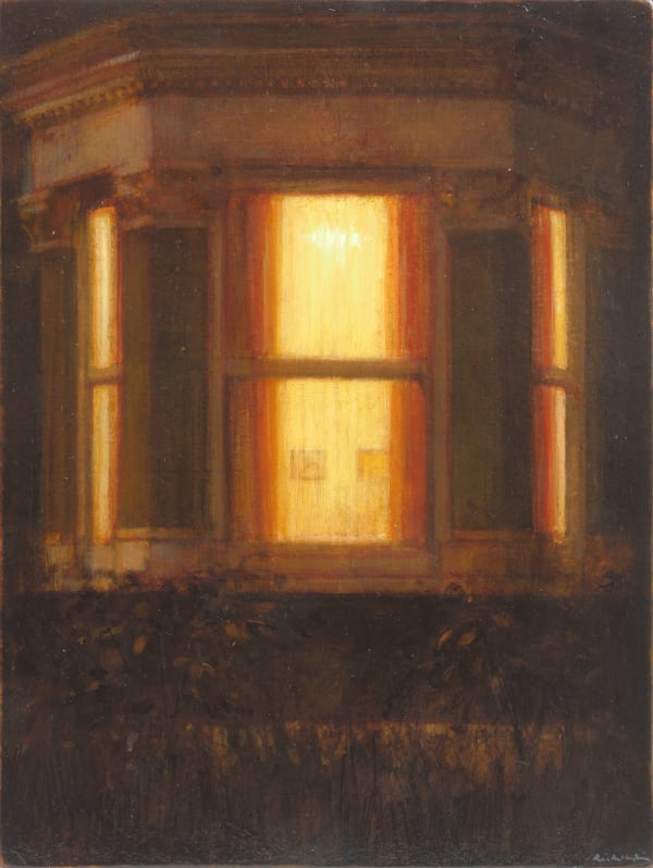 Warm yellow light emits from the interior of a house through its bay window. Plants out front barely graze the bottom of the window.