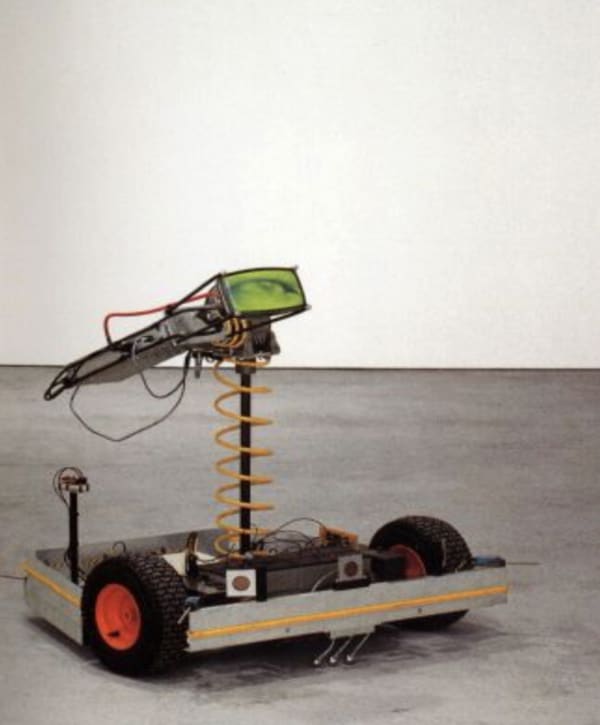 Still image of an electronic rover sculpture with two black wheels a metal boxed base custom electronics and a small elevated LCD screen. The screen displays a green colored eye