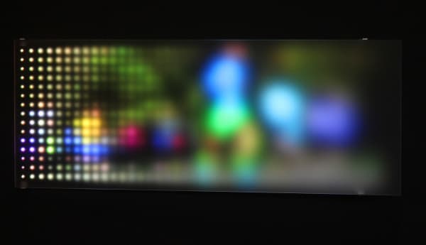Still image of a rectangular piece with multi-colored LEDs behind angled Plexiglass that display scenes of people running To the right side the scene is burry due to the Plexi being farther away while at the left is it more pixelated due to the Plexiglass being closer