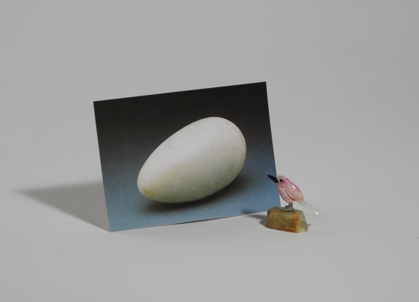 Digital duraflex photograph of a small pink bird figurine standing on a small brown stone. The bird is placed to the right of and is looking at a larger propped up photo of a white egg which is actually a popular Brancusi sculpture