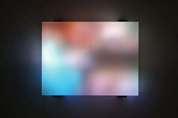 Still image of multiple multi-colored LEDs blurred behind thick block of resin show a street scene of cars and people walking