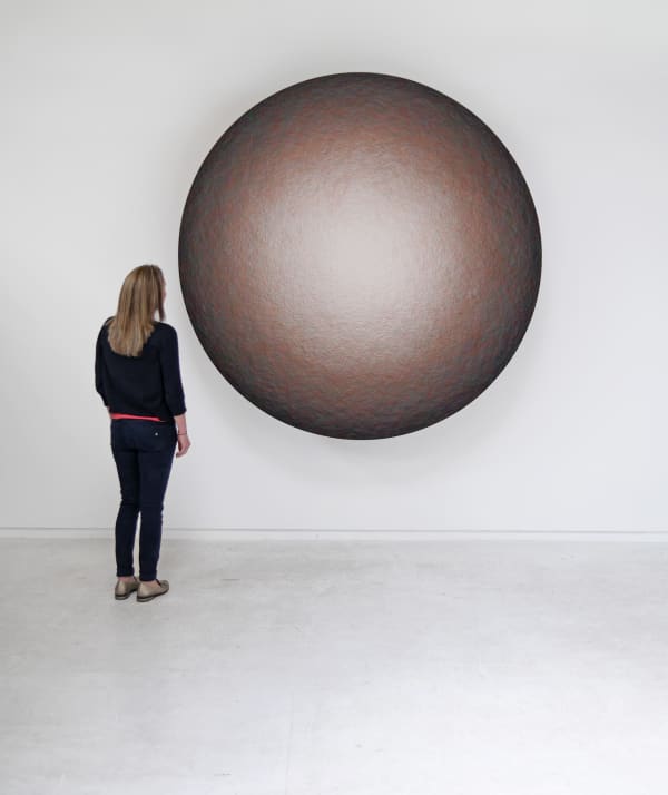 Layers of multi-colored threads strung across a round wooden and plaster panel create the effect of a glowing light brown center that is encircled by a dark brown and black border To the left of the piece a woman is present to demonstrate scale