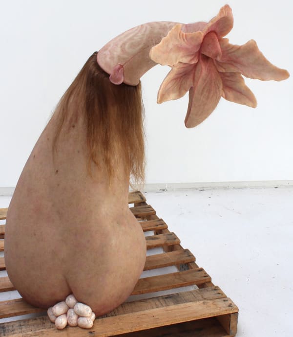 Sculpture of pear-shaped figure made of speckled human skin sits atop potato shaped eggs. The top of the figure has long brown hair and a phallic protrusion that flowers outward into six petals. Creature’ back has a tongue and a bird-like shape outlined in hair. Creature is perched on wooden planks.