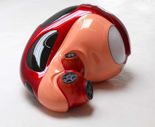Sculpture of shiny orange and red automotive figure with four wheels lying in fetal position.