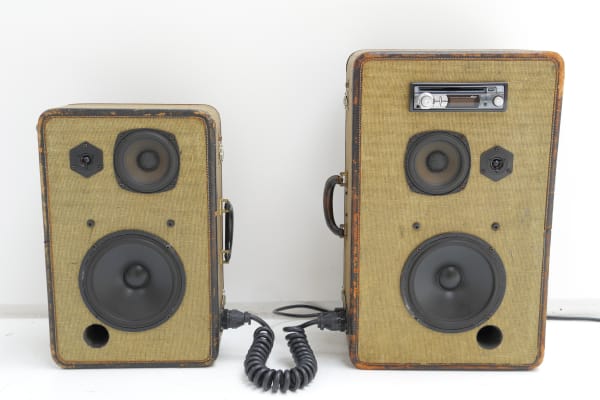 Two suitcases in two different sizes on their sides with two speakers installed on their fronts. They're connected by a coiled black cord. The larger suitcase has a stereo installed as well.
