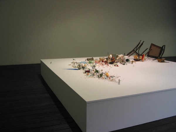 Digital photograph of an installation piece in which multiple broken and smashed objects are arranged in a curving line on a large white wooden platform. A very tiny human figurine wielding an axe above its head is placed at the front of the line and is assumed to be responsible for the damaged objects. The objects increase in size as the line curves and reaches the end of its platform