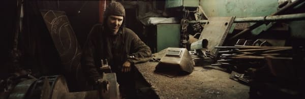 Digital panoramic portrait photograph of a Russian welder surrounded by his workbench and several tools in his darkened workshop