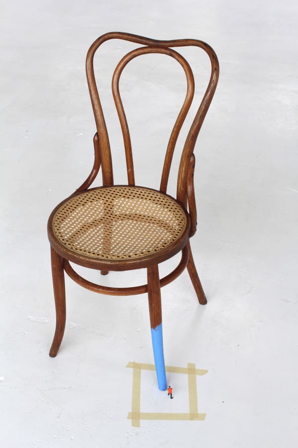 Digital photograph of an installation piece in which one half of a regular sized wooden chair is painted a bright baby blue. An incredibly tiny human figurine with a paint roller is placed next to the blue leg and is assumed to be the one responsible for painting it. Surrounding the painted leg and the tiny figurine is an uneven square of beige painters tape