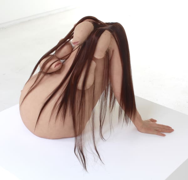 Sculpture of pregnant torso leaning back on one hand. Torso has human skin, long brown hair and many bulbous protrusions with lips on the ends.