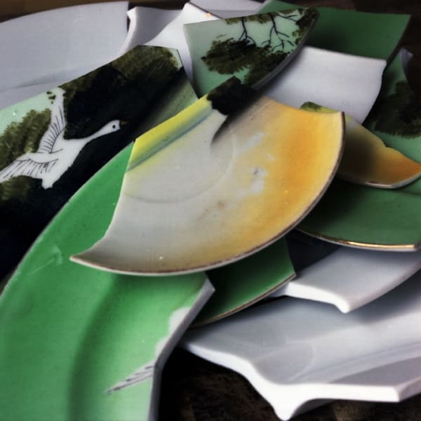 Zoomed in digital photograph of a pile of shattered yellow green and white ceramic. Painted trees and a flying white swan can be seen on some shards