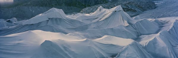 Blue toned digital panoramic landscape photograph of dirty white tarps blanketing the rough multiple ridged surface of the Swiss Rhône Glacier with rocky mountain terrain in the background
