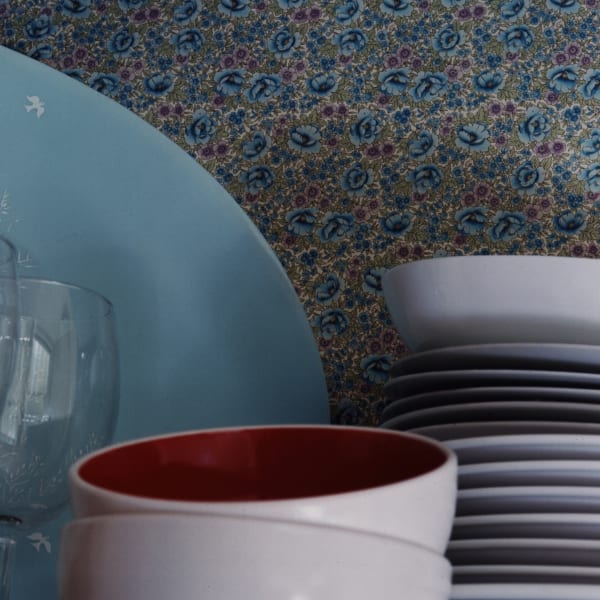 Chromogenic print of a digital photograph of a collection of multicolored pale plates, bowls, and glasses against an intricate blue green and pink floral patterned wallpaper