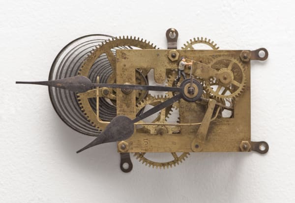 Digital photograph of an installation piece of a metallic bronze and copper gear clock. At the center of the clock on top of several gears a tiny human figurine stands and appears to be pulling on a tall lever